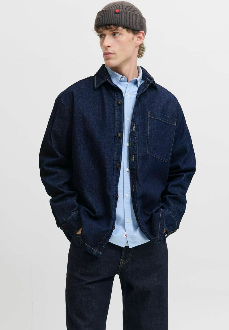 Giacca di jeans scura con chiusura a bottoni, tasca sul petto singola e vestibilità comoda, indossata sopra una camicia azzurro chiaro con bottoni. Completata con un berretto grigio.