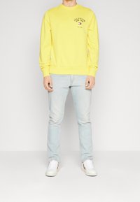 Gelbes Sweatshirt mit Rundhalsausschnitt und Logo, kombiniert mit hellblauen Jeans und weißen Sneakers. Glattes Material, lässige Passform, gerippte Bündchen.