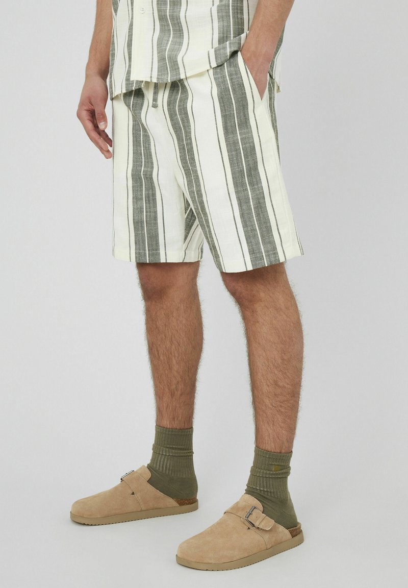 Shorts rayés en tissu blanc et gris, avec une coupe décontractée. Associés à des chaussures à enfiler beiges et des chaussettes côtelées vertes.