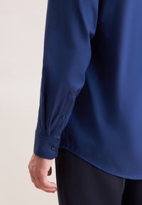 Donkerblauw overhemd met lange mouwen en een satijnen afwerking. Voorzien van een manchetknoop met twee knopen en een afgeronde zoom. Soepele textuur met een subtiele glans.