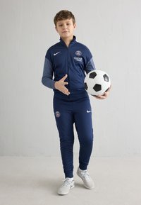 Mornarska trenirka sa svijetlosivim akcentima, s PSG logotipom i natpisom "QATAR AIRWAYS". Drži crno-bijelu nogometnu loptu, stoji na svijetlom podu.