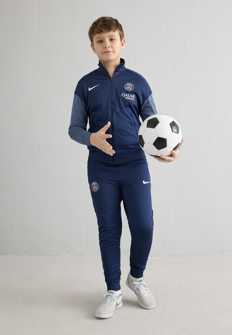 Mornarska trenirka sa svijetlosivim akcentima, s PSG logotipom i natpisom "QATAR AIRWAYS". Drži crno-bijelu nogometnu loptu, stoji na svijetlom podu.