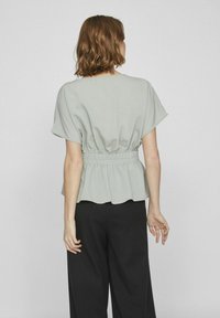 Gaiši pelēks, īsām piedurknēm tops ar pepluma vidukli, strukturēta auduma, apaļš kakla izgriezums un savilkta elastīga jostasvieta aizmugurē; saskaņots ar melnām biksēm.