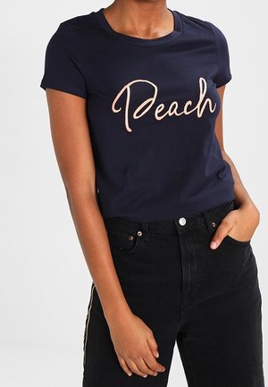 Marineblaues T-Shirt mit kurzen Ärmeln und rundem Ausschnitt, mit dem Schriftzug "Peach" in einer kursiven, hellrosa Schrift. Kombiniert mit schwarzen Jeans.