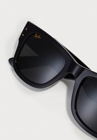 Ochelari de soare negri cu lentile întunecate, ramă pătrată îndrăzneață, logo-ul Ray-Ban auriu și detalii subtile lângă balamale. Textură din plastic neted.