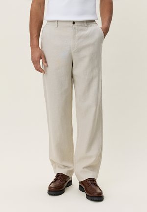 Mann trägt beige gerade geschnittene Hose und dunkelbraune Leder Schnürschuhe, steht mit einer Hand in der Tasche.