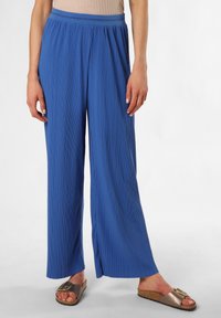 Blaue, plissierte, weit geschnittene Hosen mit elastischem Bund; leichtes Material mit strukturierter Oberfläche; getragen mit braunen Schlappen.