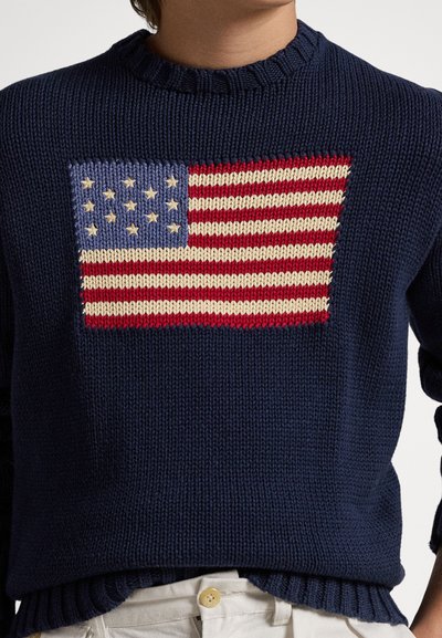 Μπλε πλεκτό πουλόβερ με μεγάλο σχέδιο της αμερικανικής σημαίας σε κόκκινο, άσπρο και μπλε. Υφασμάτινη υφή με ριμπ λαιμόκοψη και τελείωμα.