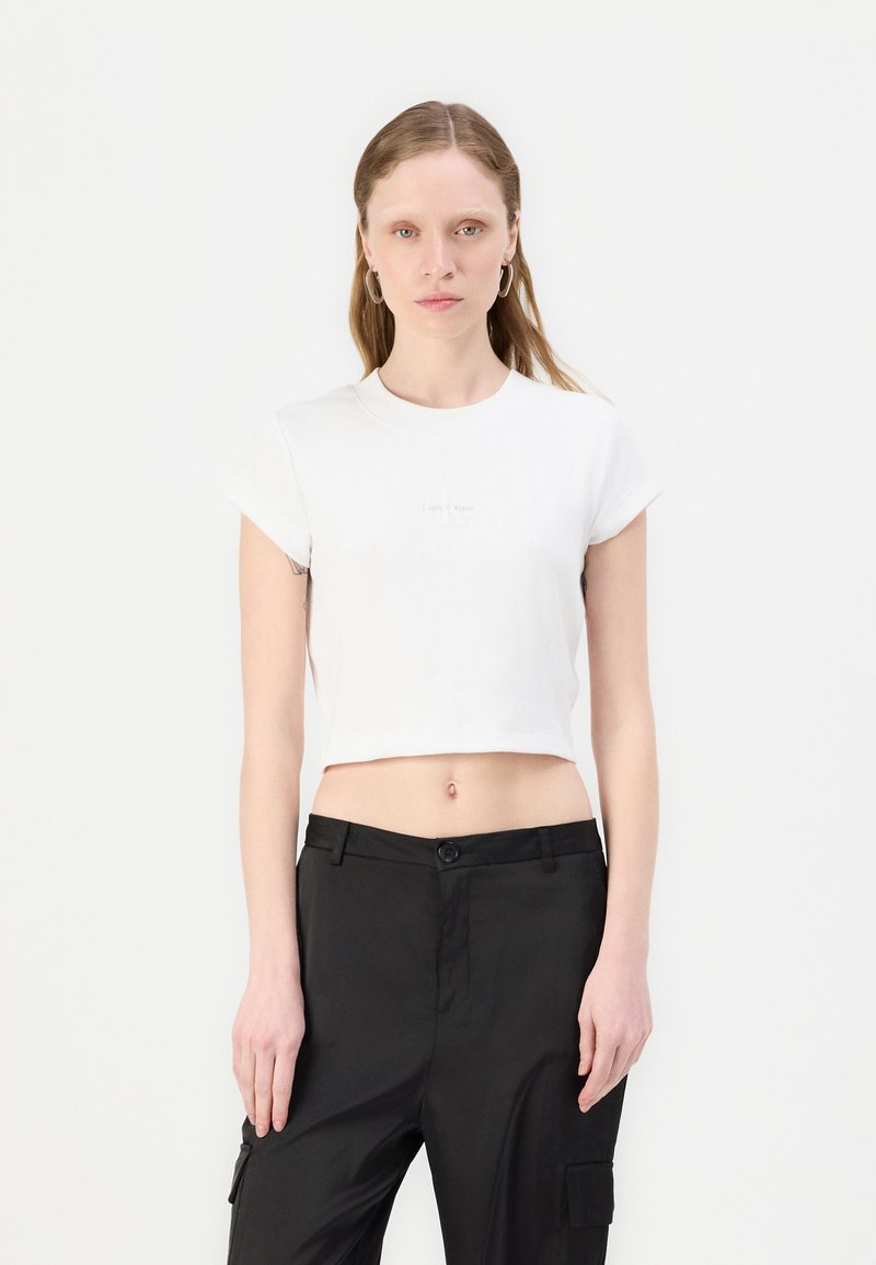 White cropped t-shirt with a fitted design, short sleeves, and a subtle logo on the chest, paired with black high-waisted pants.
