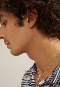 Boucle d'oreille et pendentif en argent en forme de cœur portés par un homme avec des cheveux courts et bouclés. L'arrière-plan présente une couleur neutre.