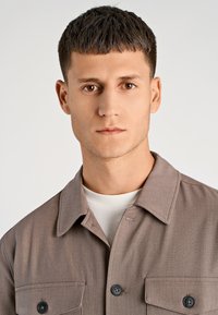 Bruine stoffen jas met twee borstzakken, knoopsluitingen en een spreidkraag, gedragen over een lichtgekleurde blouse. Soepele textuur.