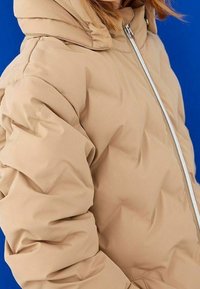 Veste matelassée beige avec capuche, présentant une texture matelassée, une fermeture éclair frontale et des poignets élastiques. Le fond est d'une couleur bleue unie.