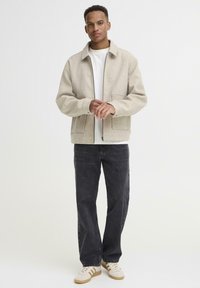 Beige, uldblandet jakke med krave, frontlommer og lynlås. Båret over en hvid skjorte med sorte denim bukser og lyse sneakers.