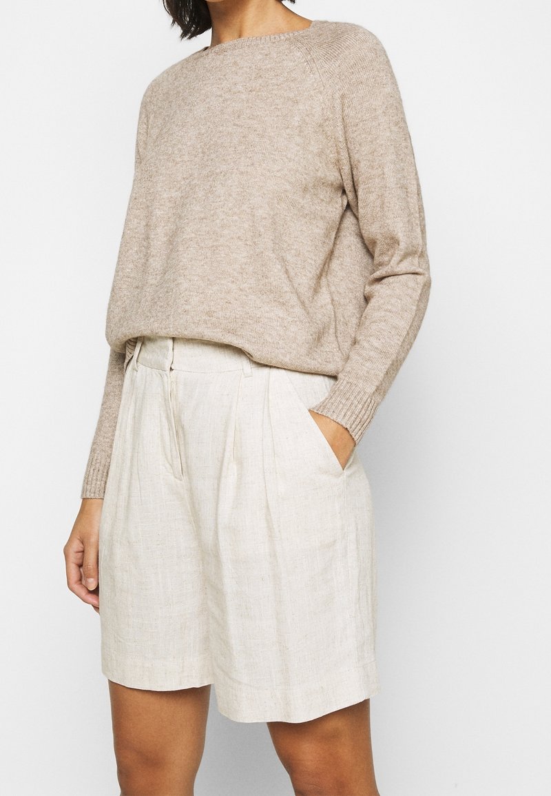 Pull beige en maille avec un col rond, porté sur un short en lin clair plissé, avec des poches latérales et une coupe décontractée.