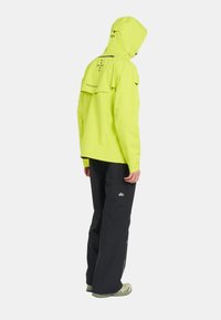 Veste imperméable jaune vif avec capuche réglable, ventilation au dos et accents noirs. Associée à un pantalon noir et des chaussures vert clair.