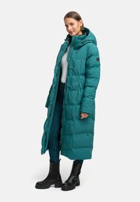 Tyrkysová puffer bunda s kapucňou, so štepovaným dizajnom, gombíkmi a bočnými vreckami. Nosená cez biele tričko a džínsy s čiernymi topánkami.