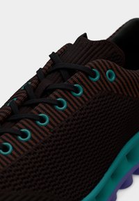 Κοντινό πλάνο μαύρου και καφέ ριγωτού αθλητικού παπουτσιού με μαύρα κορδόνια, τυρκουάζ οπές και λεπτομέρειες στη σόλα σε τυρκουάζ και μωβ.