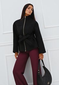 Giacca nera con zip, vita aderente, dettaglio della cintura a fiocco e maniche ampie. Abbinata a pantaloni bordeaux a righe e una borsa nera trapuntata.
