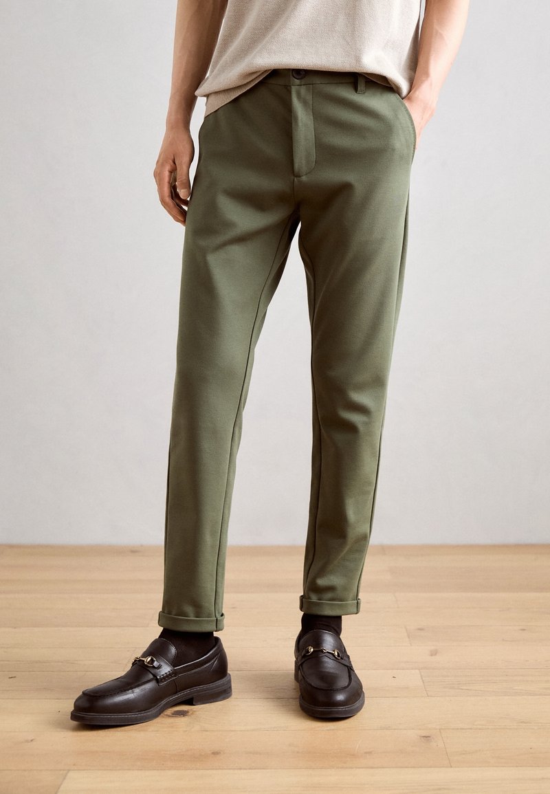 Corps inférieur portant un pantalon slim vert olive, des mocassins marron avec détail de boucle en métal, et un haut beige, debout sur un sol en bois.