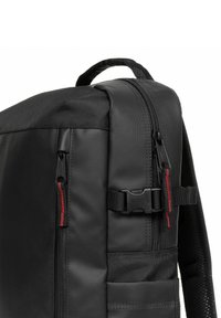 Černý batoh s texturovanou látkou, zipy se červenými akcenty a boční kapsou. Obsahuje pevnou rukojeť a nastavitelné popruhy.