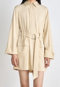 Robe chemise beige ceinturée avec un col, des manches longues, des poches latérales et une texture lisse. Présente un logo discret et une coupe structurée.