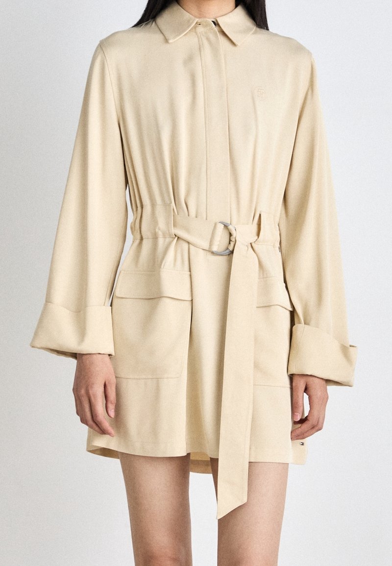 Robe chemise beige ceinturée avec un col, des manches longues, des poches latérales et une texture lisse. Présente un logo discret et une coupe structurée.