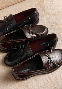 Două perechi de mocasini: una din piele neagră cu fundă decorativă și una maro cu model de crocodil și fundă asortată. Ambele au tălpi interioare căptușite.