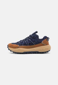 Marine- und braune Trail-Schuhe mit strukturiertem Obermaterial, verstärkten Überzügen und einer auffälligen gepolsterten Sohle mit Profilstollen. Rote Markenakzente.