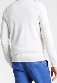 Homme portant un pull blanc uni à manches longues et un pantalon bleu, vu de dos, debout devant un fond blanc.