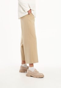 Pantaloni beige a gamba larga con una texture liscia, abbinati a mocassini beige spessi e calzini bianchi, visti di lato.