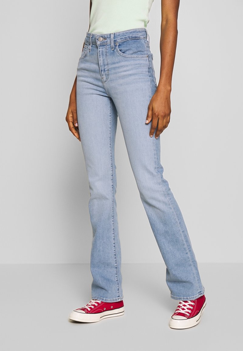 Personne portant un jean droit bleu clair, un top blanc et des baskets en toile rouges, debout devant un fond clair uni.