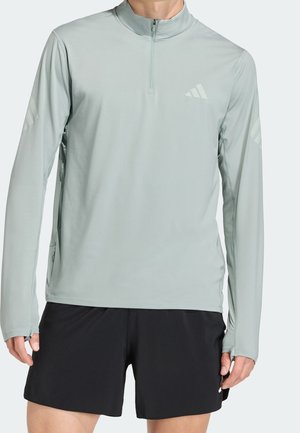 Man draagt een lichtgrijze atletische top met lange mouwen en een rits tot een kwart, en zwarte korte broek, staand met de armen ontspannen langs het lichaam.