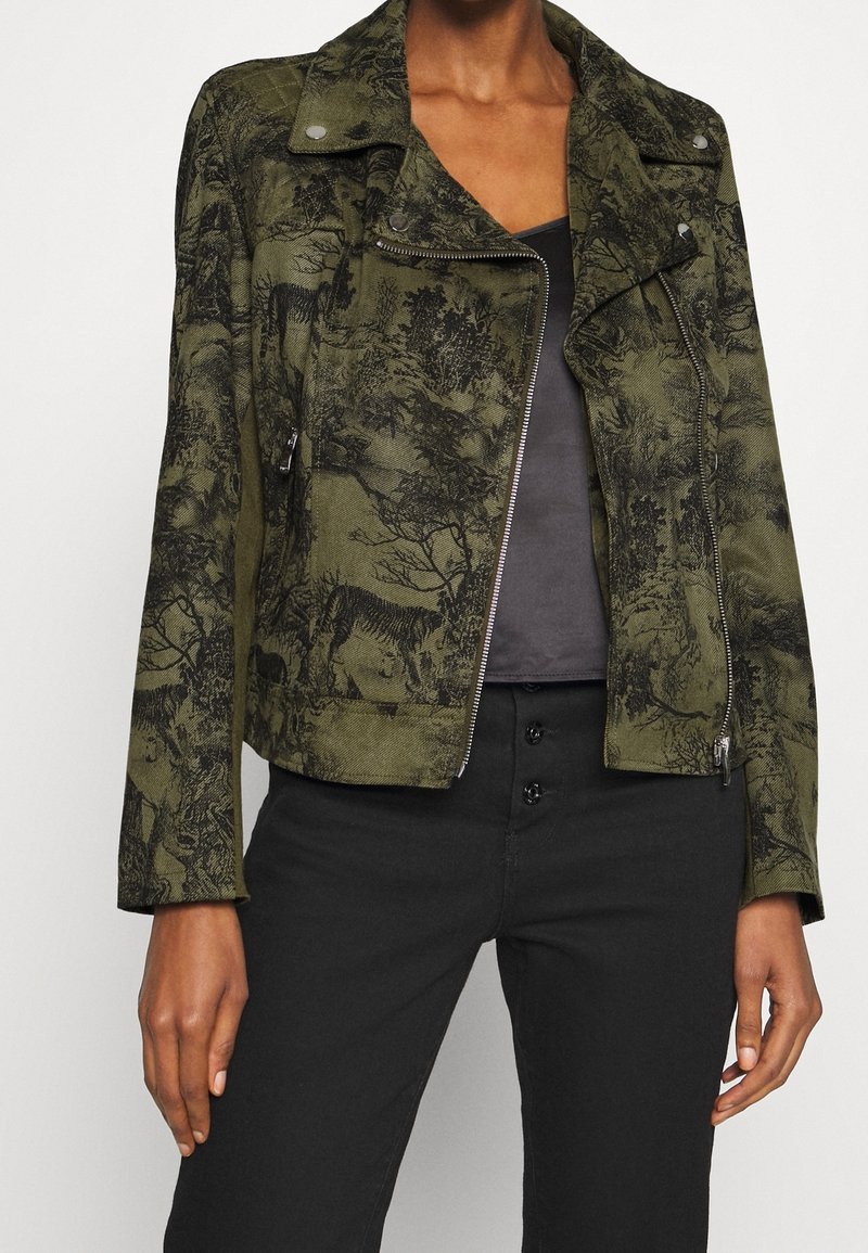 Veste verte zippée avec imprimés noirs de nature et d'animaux portée sur une chemise sombre, associée à un pantalon noir taille haute avec boutons.