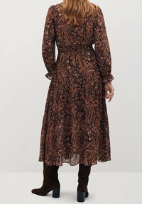 Robe longue à fleurs sombres avec un motif paisley aux teintes chaudes, à manches longues, corsage cintré et jupe fluide. Portée avec des bottes marron.