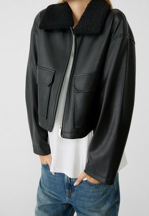 Vrouw draagt zwarte cropped leren jas met shearling kraag, wit overhemd en blauwe jeans, handen in de zakken, staand tegen een effen achtergrond.