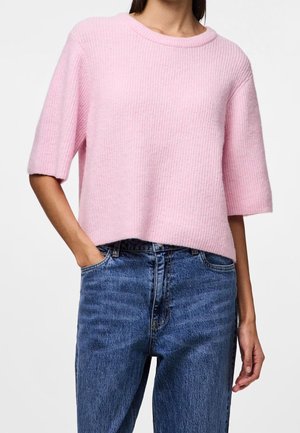 Kvinde iført en løs, lys pink ribbestrikket sweater med ærmer til albuen og højtaljede blå jeans, hånd i lommen.