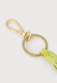 Złoty metalowy brelok z grawerem "COCCINELLE", przymocowany do limonkowozielonego skórzanego frędzla i złotego karabińczyka na białym tle.