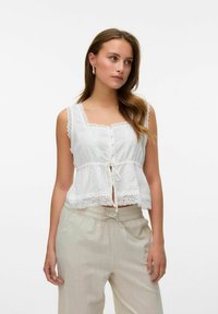 Haut sans manches blanc avec un bord en dentelle, des fermetures à boutons et un détail de ceinture à nouer. Assorti à un pantalon clair et ample.