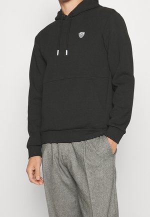 Sudadera negra con capucha, bolsillo frontal tipo canguro, cordones y un logo en el pecho. La tela parece suave y lisa. Combinada con pantalones grises de patrón.