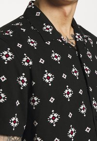 Chemise noire à manches courtes avec un motif géométrique en blanc et rouge, dotée d'un col classique et d'une texture de tissu douce.