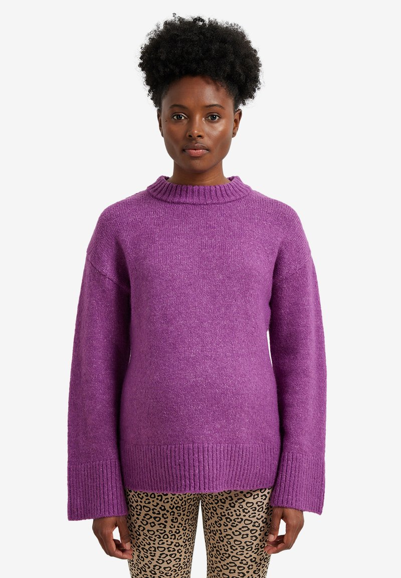 Frau mit lockigem Haar, die einen lila gestrickten Pullover und eine Hose mit Leopardenmuster trägt, steht vor einem schlichten weißen Hintergrund.