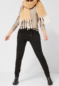 Un pantalon de survêtement noir avec une taille élastique et un cordon de serrage, associé à un haut à manches longues marron clair et à une écharpe orange à motifs avec des franges.