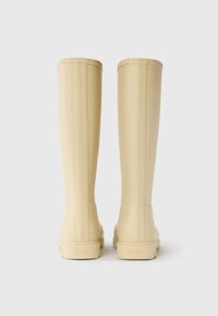 Beige rubber rain boots with a smooth texture, tall shaft, and ridged accents; featuring a logo on the heel and a rounded toe design.