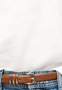 Καφέ δερμάτινη ζώνη με χρυσή αγκράφα, φορεμένη με ανοιχτό μπλε τζιν παντελόνι και λευκό υφασμάτινο τοπ.