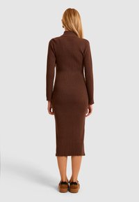 Abito marrone a coste con maniche lunghe e una silhouette aderente, che arriva a metà polpaccio; indossato con scarpe con piattaforma che mostrano un design testurizzato.