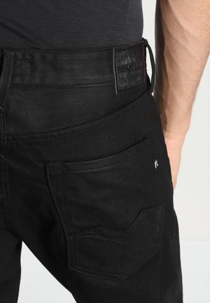 Gros plan sur une personne portant un jean noir avec une poche arrière et une étiquette de marque en cuir visible près de la ceinture.