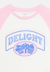 T-shirt raglan blanc et rose avec le texte "DELIGHT" et une illustration de trois cupcakes décorés de cœurs sur le devant.