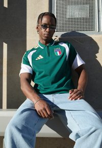 Πράσινο αθλητικό πόλο μπλουζάκι με λευκές λεπτομέρειες και χρυσές λεπτομέρειες, με διακριτικό Ιταλίας. Φοριέται με ανοιχτό μπλε τζιν, ομαλή υφή, άνετη εφαρμογή.