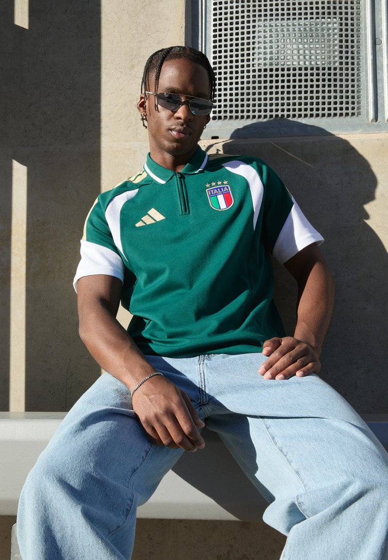 Πράσινο αθλητικό πόλο μπλουζάκι με λευκές λεπτομέρειες και χρυσές λεπτομέρειες, με διακριτικό Ιταλίας. Φοριέται με ανοιχτό μπλε τζιν, ομαλή υφή, άνετη εφαρμογή.