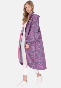 Lång lila regnrock med rosa leopardmönster, utrustad med huva, elastiska ärmslut och dragkedja framtill. Bärs över vita byxor och sneakers.